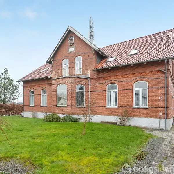 Rødt murstenshus med buede vinduer og rødt tegltag står i en grøn have med træer. Husnummeret "17" er synligt på væggen. Grussti leder op til huset.