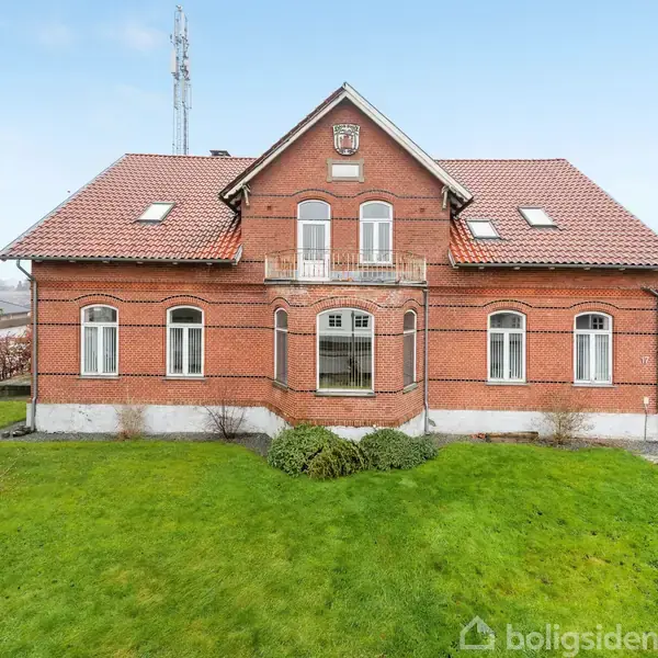 Et rødstenet hus med rødt tegltag står i en velplejet have. Huset har fire store vinduer og en centralt placeret dør. Omkranset af grønt græs og hække.