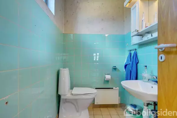 Toilet i aktion i et lille badeværelse med mintgrønne fliser; en vask med spejl hænger til højre. Der er et håndklæde og hylder med toiletartikler.