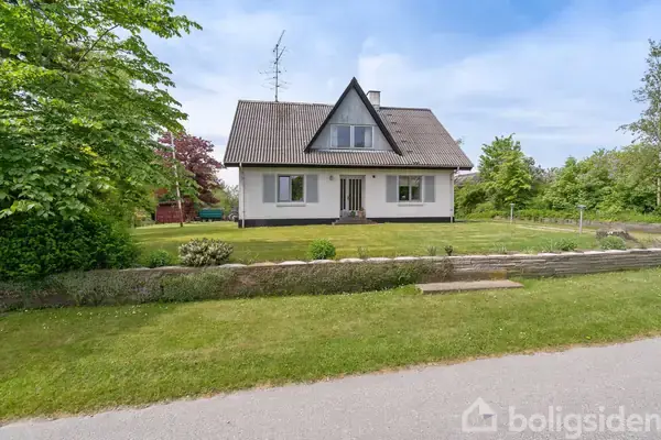 Hus står på grøn græsplæne, omgivet af træer og buske. Det har en trekantet gavl, gråt tag og hvide vægge. En sti fører op til hoveddøren.