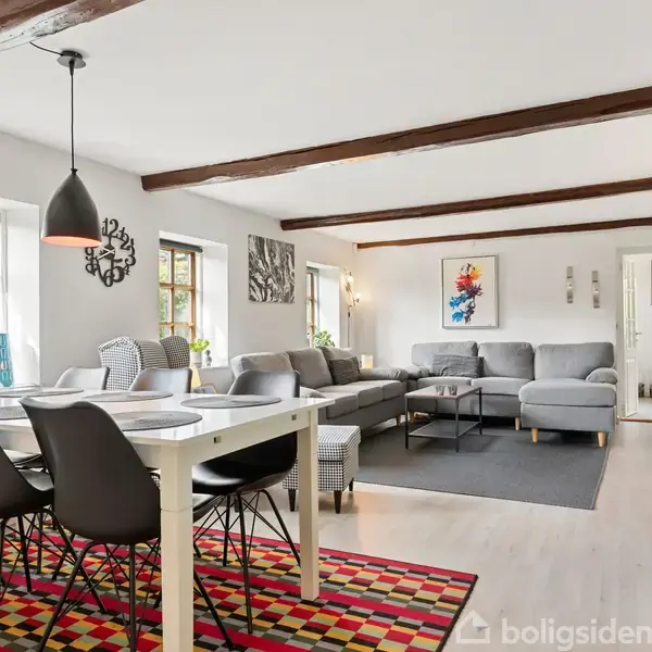 Der er et lyst og rummeligt stueareal med spisebord og stole til venstre, en stor grå hjørnesofa til højre, dekoreret med farverige tæpper og vægkunst.