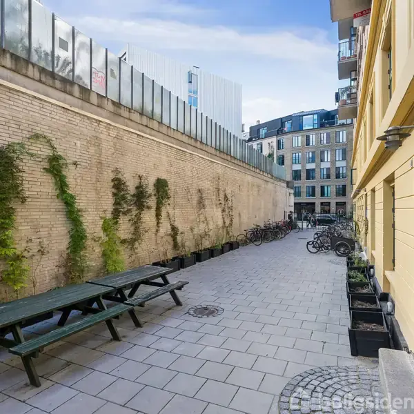 En bænk står stille i en smal gård, omgivet af vægge med klatreplanter og cykler. Moderne bygninger tårner i baggrunden under en blå himmel.