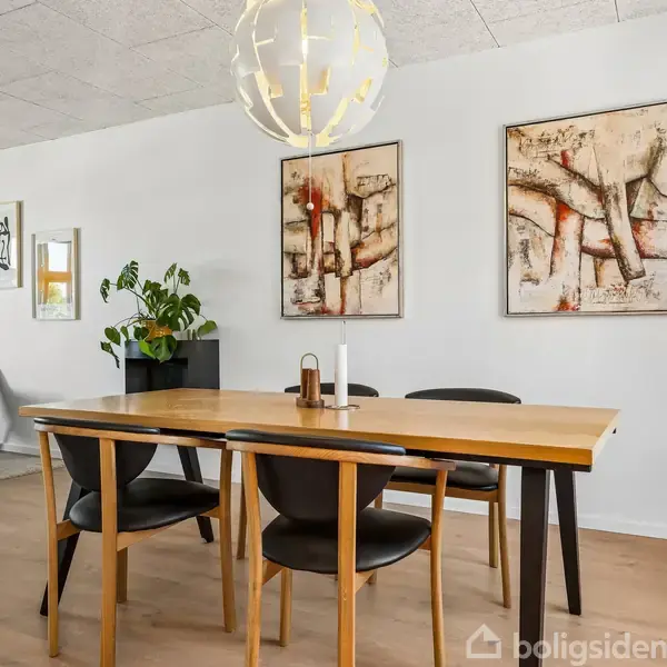 Spisebord med fire stole står i en moderne stue. Over bordet hænger en rund loftlampe, og på væggene er der abstrakte malerier. Planter og kunst pryder rummet.