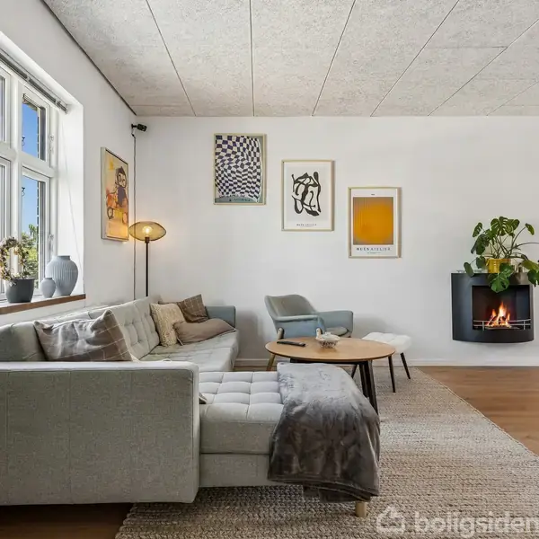 Grå sofa står i lys stue med trægulv og vægkunst. Rundt sofabord foran, lænestol ved siden af. Pejs på væggen med planter ovenpå. Sollys kommer ind fra vinduerne.