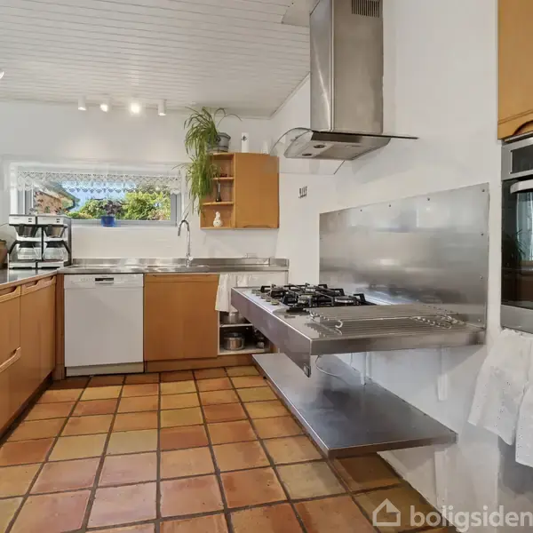 Et moderne køkken med træskabe har en indbygget ovn, gaskomfur og køkkenø. Vinduer lukker lys ind og planter dekorerer rummet. Flisegulv og hvidt loft skaber en lys atmosfære.