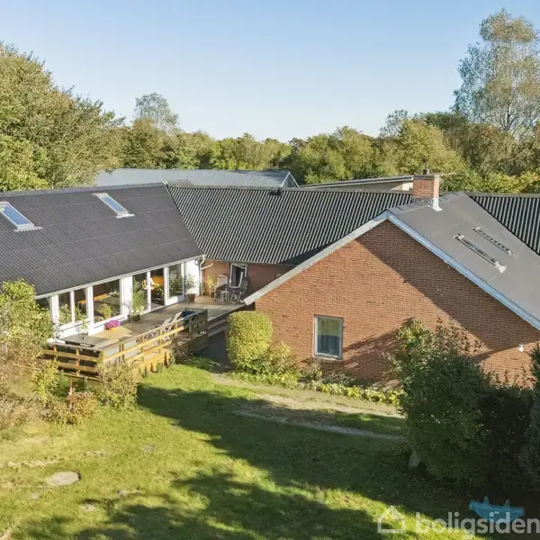 Hus med sort tage og røde murstensvægge ses fra en skrå vinkel. Det ligger i en have omkranset af træer og buske, hvilket giver en naturlig, fredfyldt atmosfære.