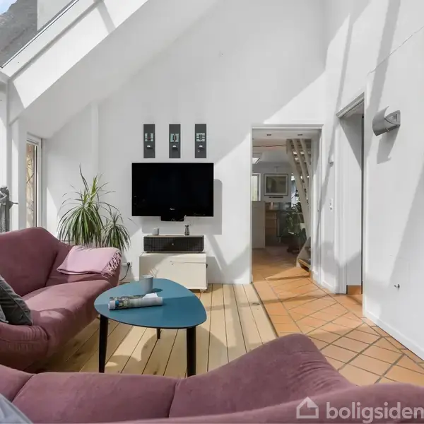 Stue med sofaer i lilla, en fladskærms-TV på hvid væg, og et lille blå bord. Store vinduer giver naturligt lys. Der er adgange til andre rum og dekorationer på væggen.