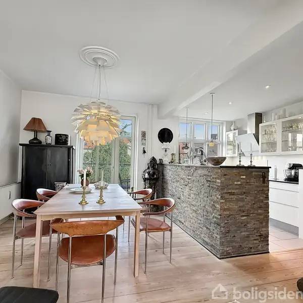 Spisebord med seks stole omgivet af en indbydende spisestue. Bordet har tre lysestager, og rummet har en moderne lampe. Køkkenet i baggrunden har hvide skabe og en bar med stenbeklædning.