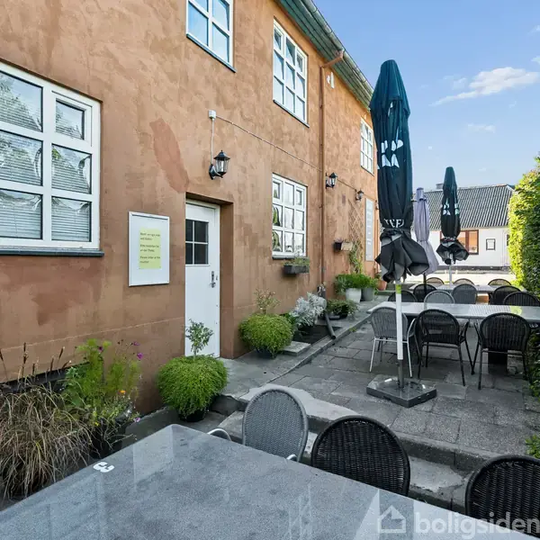 Terrassemøbler står på en flisebelagt gårdhave ved en gul bygning med vinduer. Flere parasoller er foldet sammen. En dør med et skilt er synlig. Grønne planter omgiver området.