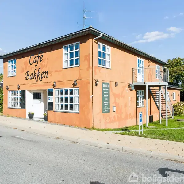 Bygning med orangemalede vægge og skiltet “Cafe Bakken”. Trappe på siden, omgivet af grønne buske, gade og fortov under en blå himmel.