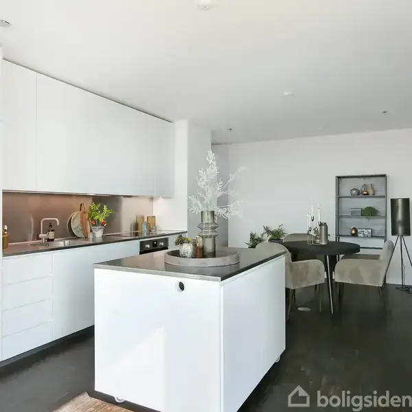 Et moderne køkken-alrum med hvidt køkken, ø i forgrunden, og spisebord i baggrund. Lysindfald fra oven, vægdekorationer og hylde med planter og bøger skaber en indbydende atmosfære.