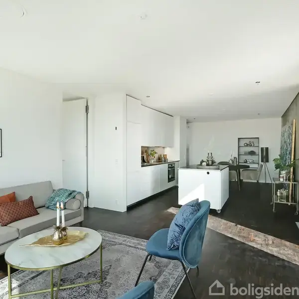 Stue med grå sofa og blå stole, placeret ved et rundt sofabord. Åbent køkken ved siden af, med hvid køkkenø. Store vinduer til højre slipper lys ind.