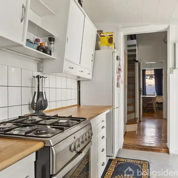 Komfur med gasblus står i et smalt køkkenrum. Køkkenet har hvide skabe, flisevæg og træbordplader. En åben dør fører til en gang mod en spisestue. En airfryer står på bordet.
