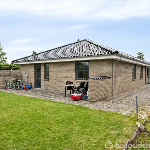 En lav, firkantet murstensbygning med mørkt tegltag står på en flisebelagt gårdsplads. Ved siden af bygningen er der små buske, og en træhegn adskiller ejendommen fra naboen.