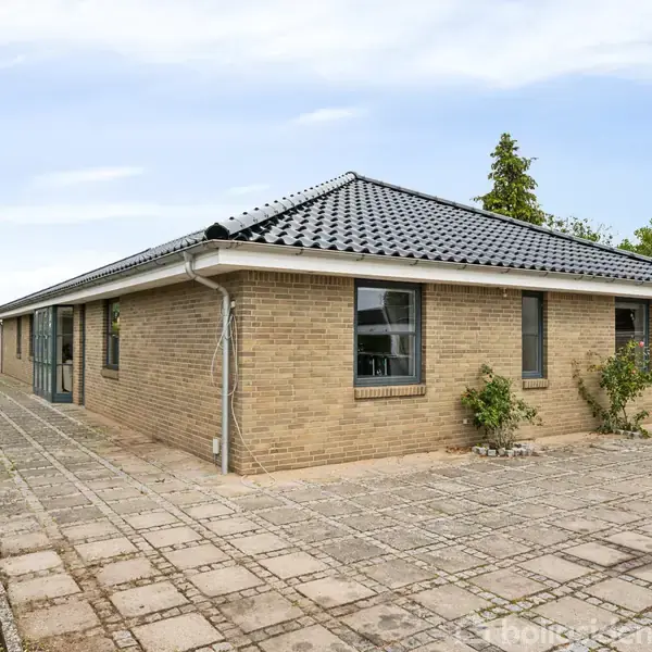 En etplans villa med murstensfacade og sort tegltag står på en flisebelagt indkørsel. I baggrunden ses en grøn have og et træhus.