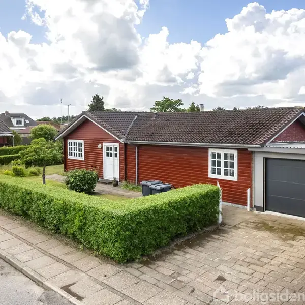 Rødt hus med hvide vinduer, står stille langs en stille vej. Det er omgivet af en velplejet hæk og en tilknyttet garage med en indkørsel.