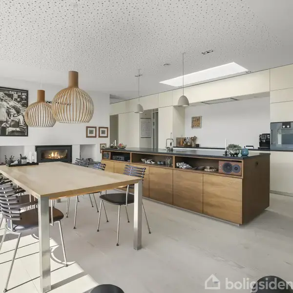 Et moderne køkken med træspisebord og metalstole, omgivet af minimalistiske hvide vægge. Hængelamper i naturmateriale hænger over bordet, og der ses en pejs i baggrunden.