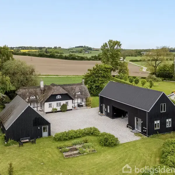Et bindingsværkshus med stråtag står stille, omgivet af landlig idyl med grønne marker og træer. To sorte træbygninger ligger på gårdspladsen, der er omgivet af græsplæne.