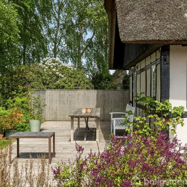 En træterrasse med bord og stole står op ad et bindingsværkshus. Blomster og buske omgiver området, og der er frodig vegetation i baggrunden.