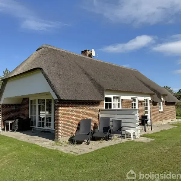 Spisebord med seks stole står i solrigt alrum med hvide og pink loftsbjælker, omgivet af store vinduer. Køkkenhjørne med mikroovn og kogeplader ses i baggrunden. Udsigt til græsplæne udenfor.