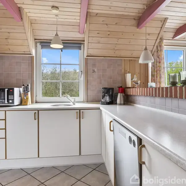 Køkken med hvide skabe, mikroovn og opvaskemaskine, beliggende under et skråt træloft. Belysning hænger fra loftet over en lys bordplade. Vinduer viser grønne omgivelser udenfor.