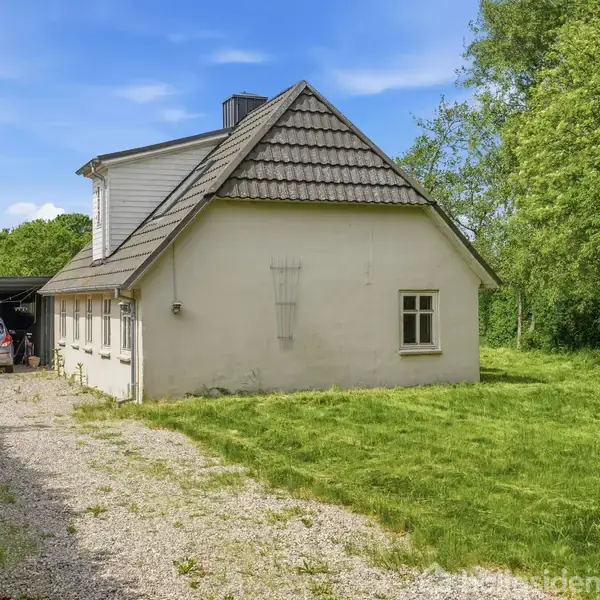 Et hvidt hus står stille ved en grusvej i en grøn, græsklædt have omgivet af træer. En bil holder under en carport i baggrunden.