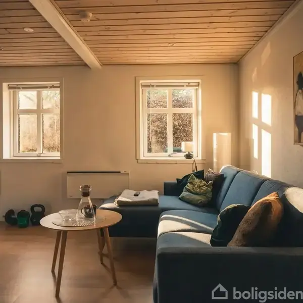 En blå sofa står i et lyst og hyggeligt rum med trægulv. To vinduer lader sollys strømme ind, og en rund træbord holder en karaffel. Væggene er dekoreret med kunst.