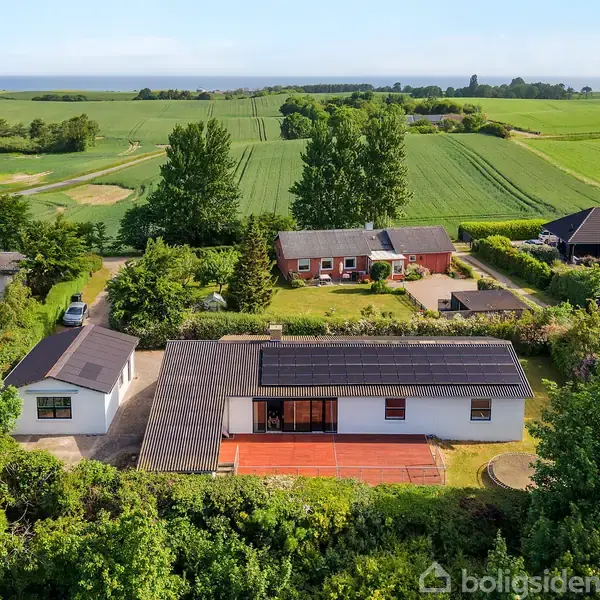Et hvidt hus med solpaneler på taget står i forgrunden omgivet af have. Baggrunden består af åbne, grønne marker og spredte træer under en klar himmel.
