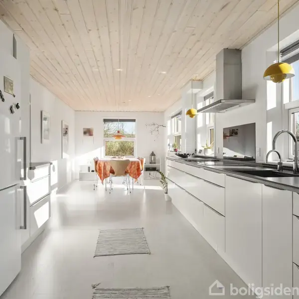 Et moderne køkken med hvide skabe langs væggene; sollys strømmer ind fra store vinduer. Et spisebord med orange dug står i baggrunden. Loftet har en lys træfinish.