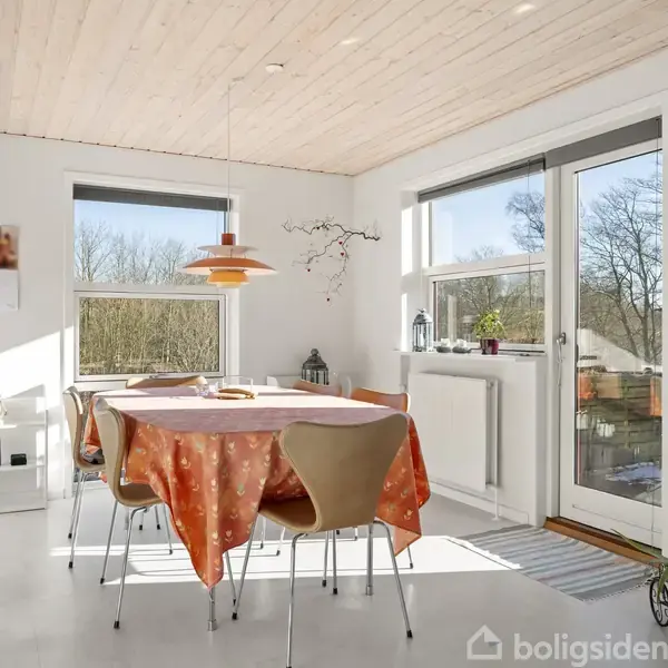 Et spisebord dækket med en orange dug står i et lyst rum med store vinduer; omgivet af stole, planter og en lampe. Udenfor ses træer gennem det soloplyste glas.