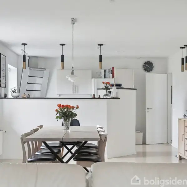 Et spisebord med blomster i vase står i en moderne, lys stue-køkken. Der er stole omkring bordet. Bagved ses et åbent køkken med hvide skabe og en trappe. En væghængt ur findes også.