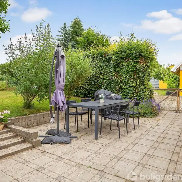 Et udendørs terrasseområde med et sort bord og flere stole. En parasol står ved siden af. Området er omgivet af grønne planter og en gul husfacade i baggrunden.