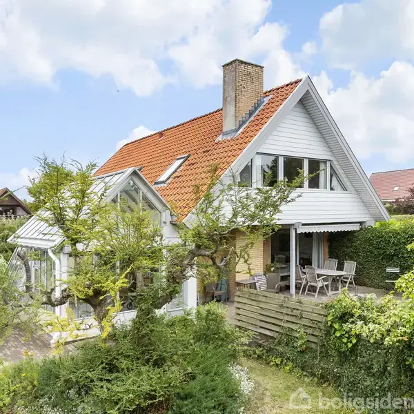 Et hvidt hus med rødt tag står omgivet af en frodig have. Terrasse med havemøbler ses foran huset. Skyer og blå himmel præger baggrunden.