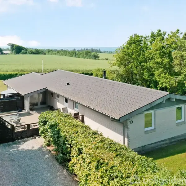 En lang, lys bygning med grå tag ses fra oven, omgivet af grønne træer og marker. Bygningen har terrasser og en grusvej. Husnummer 10 er synligt på facaden.