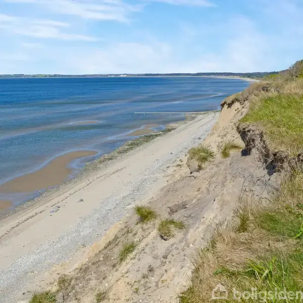 Klippekysten strækker sig langs en bred strand, hvor græs og buske vokser øverst, mens havet roligt skyller ind mod kysten under en klar, blå himmel.