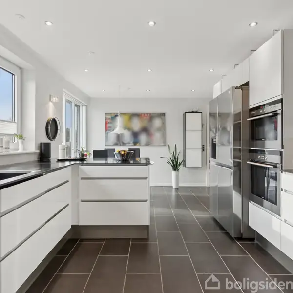 Et moderne køkken med hvide skabe og sorte bordplader står oplyst af flere vinduer. Køkkenmaskiner og krydderier er placeret på bordet. Et maleri hænger på væggen i baggrunden.