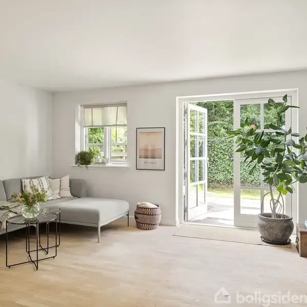 Sofa med puder står i stuen ved et lille bord; rummet er lyst med en åben dør til en terrasse og store planter ved siden af. En kurv står nær vinduet.
