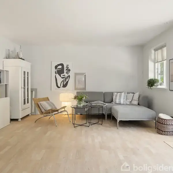 Stue med grå hjørnesofa, pyntepuder og et lavt rundt sofabord; omgivet af lyse træmøbler, reol med bøger og kunst på væggen. Store vinduer giver naturligt lys.