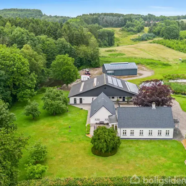Bygning ligger i frodig natur, omgivet af skove og marker. Strukturen består af flere sammenhængende bygninger med skrå tage, placeret i velplejet have med græsplæne og træer.