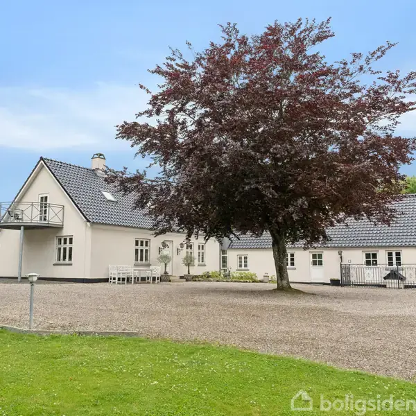 En hvid bygning med sort tag står stille, omgivet af en grusplads og grønne træer. En stor træ i forgrunden og en blå himmel bagved.