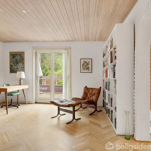 Stue med trægulv har et skrivebord, en lænestol og en bogreol. Store vinduer åbner ud mod en terrasse med udsigt til grønne omgivelser, og kunst hænger på væggene.