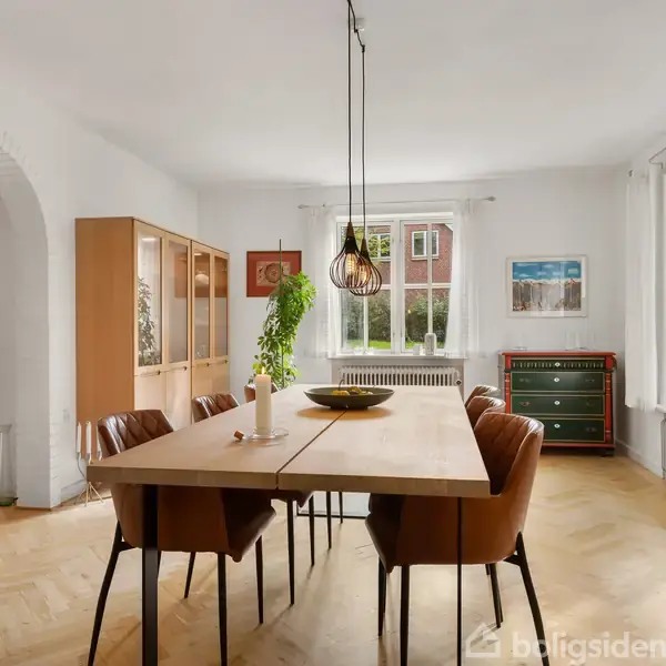 Et rektangulært spisebord i træ med brune stole står centralt. Et moderne lysekrone hænger over bordet. Rummet har lyse trægulve, store vinduer med udsigt og grønne planter.