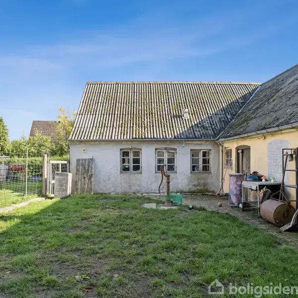 Et gammelt, gulmalet hus står stille i en solrig have med græsplæne. Ved siden af huset er et hegn, forskellige genstande og udstyr opbevaret langs muren.