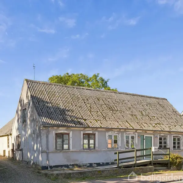 En ældre, hvid malet bygning med skråtag og små, rektangulære vinduer står stille i et solrigt kvarter. En klar blå himmel fylder baggrunden.