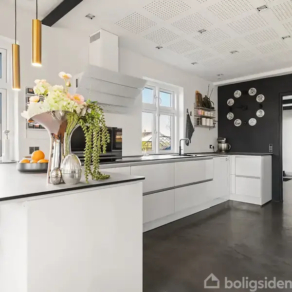 Et moderne køkken med hvide skabe og en sort bordplade. En vase med blomster står på øen. Store vinduer giver naturligt lys; væg med dekorative tallerkener.
