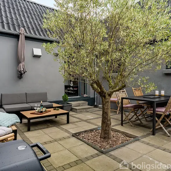 Terrasse med træ i midten omgivet af møbler; en grå sofa med sofabord til venstre, et spisebord med stole til højre, gråt plæneflisegulv og en grill i forgrunden.