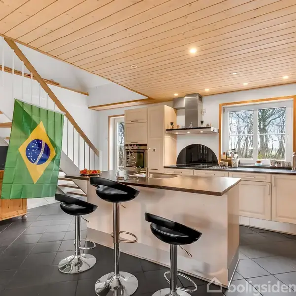 Et moderne køkken med en central ø med tre sorte barstole. En brasiliansk flag hænger ved en trappe. I baggrunden ses træbeklædt loft og vindue med udsigt til træer.