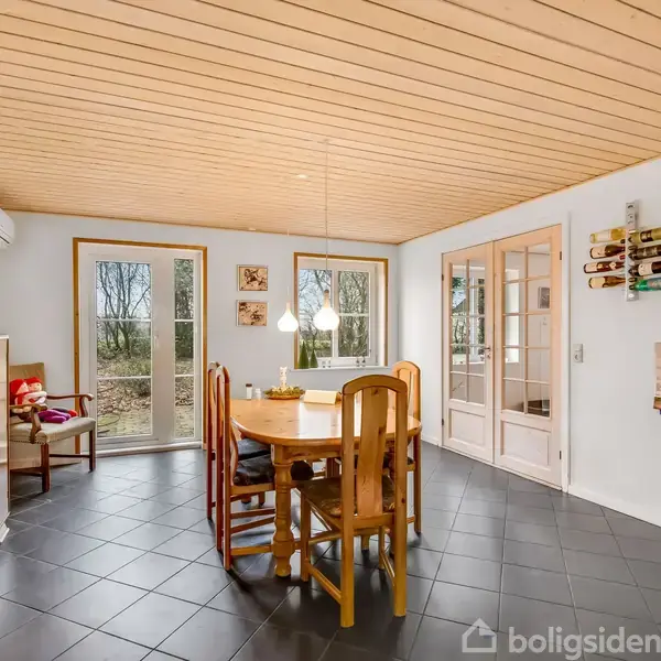 Et rundt spisebord med træstole står i et lyst rum. Væggene har vinflasker monteret, og store vinduer viser et udvendigt skovområde. Gulvet har mørke fliser.