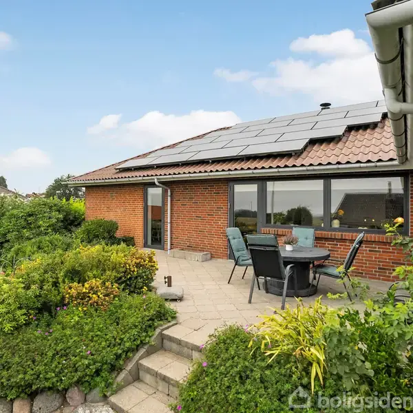 Rødt murstenshus med solpaneler på taget har en terrasse med havemøbler. Det er omgivet af en velplejet have med grønne buske og græsplæne under en blå himmel.