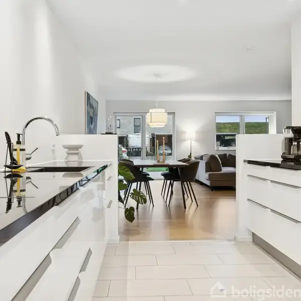 Et moderne køkken med sorte bordplader og hvide skabe fører til en lys stue med spisebord, sofa og store vinduer. En kaffemaskine og en plante ses på køkkenbordet.